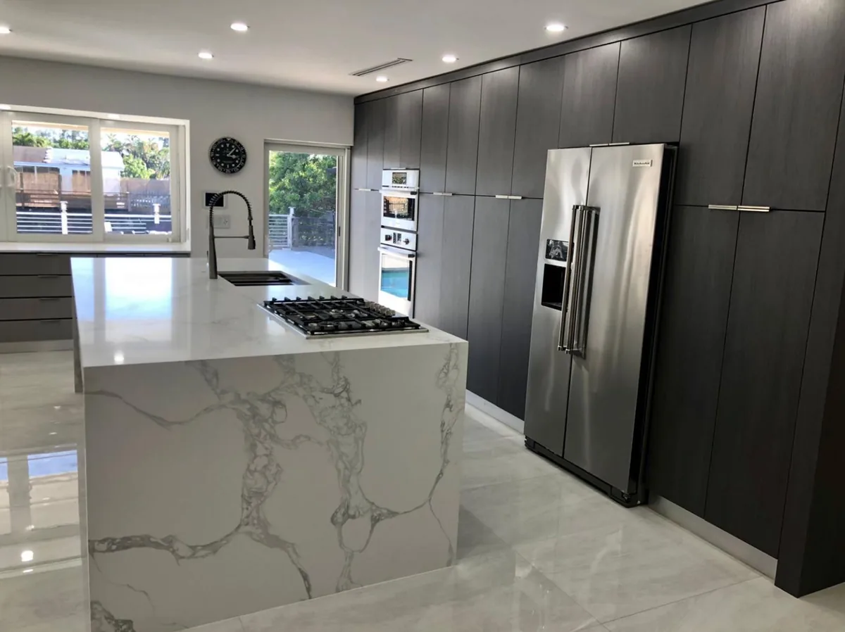 Tall dark cabinet wall with built in refrigerator facing a white quartz waterfall island with cooktop