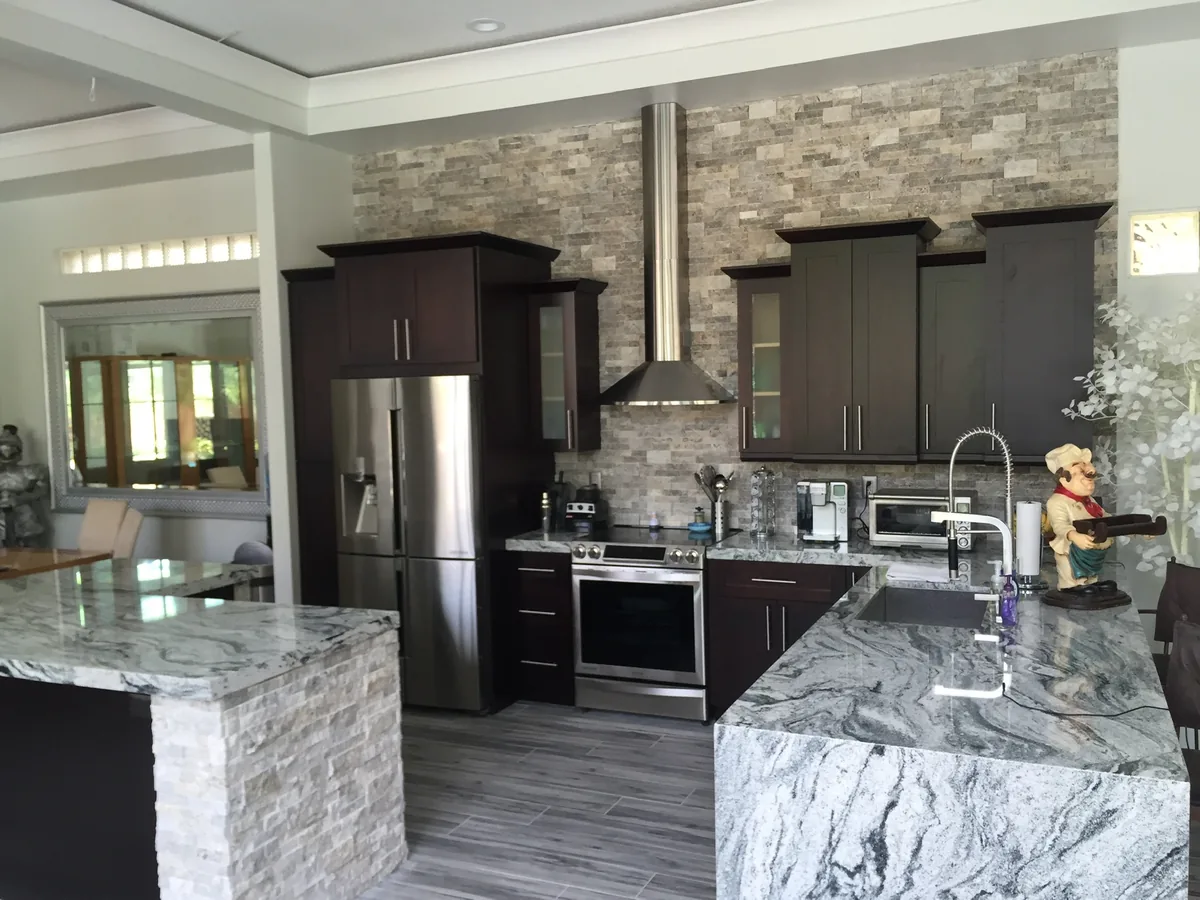 Espresso kitchen with stainless chimney hood, stacked stone wall and granite waterfall peninsula