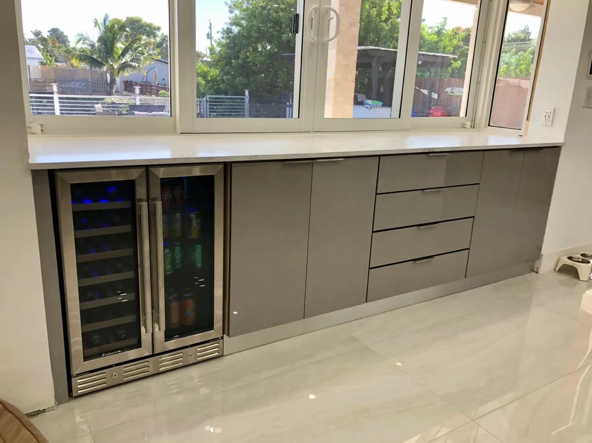 Gray flat panel base cabinets with a dual zone wine fridge under a window and white quartz counter